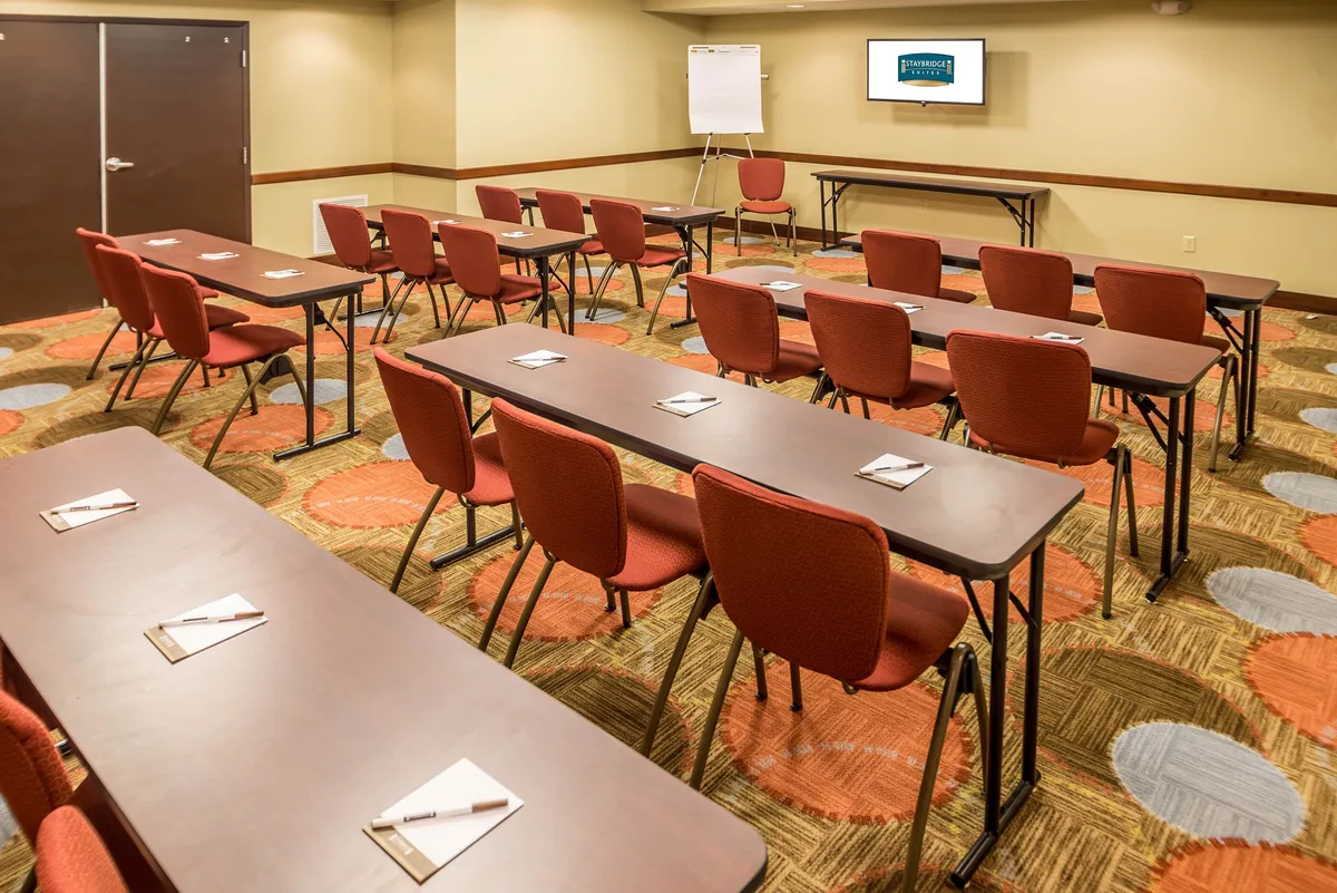 A flexible lobby or event space is shown with tables arranged in rows and a presentation screen at the front, evoking a welcoming venue for the evening reception or social gatherings. The setup is versatile, ideal for both business-focused and relaxing events after a day of activities.