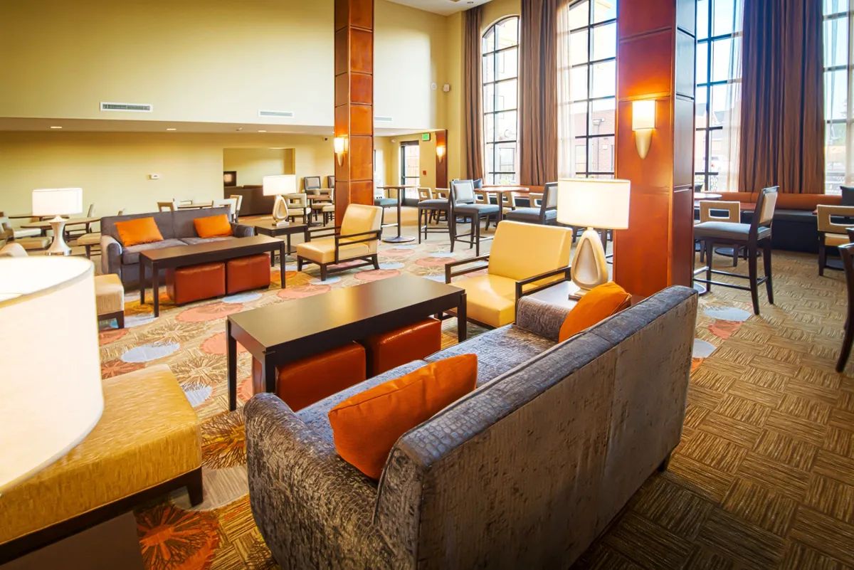 A spacious living area with large windows letting in abundant natural light, featuring comfortable seating, a decorative carpet, and stylish lighting fixtures. The space is arranged for both relaxation and socializing, with a cozy atmosphere suitable for reading or casual dining.