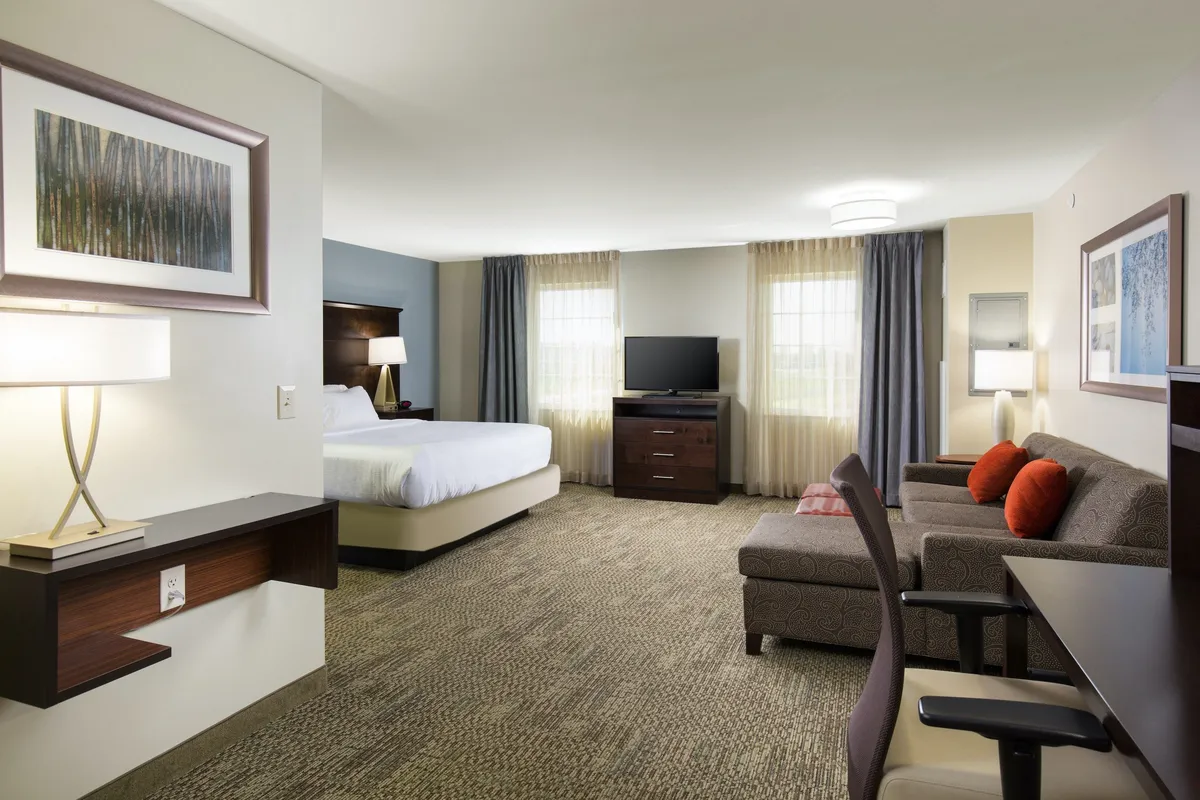 The image showcases a modern, inviting suite with a plush king bed, comfortable sofa, soft lighting from a stylish lamp, and a flat-screen television—creating a warm and cozy atmosphere ideal for relaxing after exploring Denver. The setting suggests ample space for unwinding, reading, or enjoying a movie night, all within a thoughtfully designed and welcoming interior.