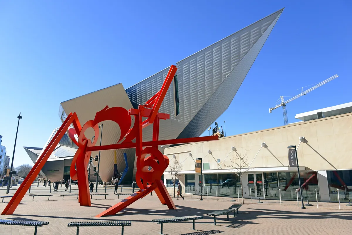 This image spotlights a striking red outdoor sculpture set against the backdrop of a modern architectural building, representing Denver’s thriving arts scene. People gather nearby, suggesting the area is a hub for social interaction, photography, and cultural appreciation.