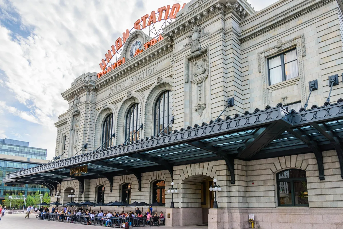 This vibrant exterior shot features Denver's historic Union Station with its grand clock, architectural detailing, and the glowing neon 'UNION STATION' sign. The foreground showcases bustling outdoor cafe seating, inviting visitors to relax and soak up the local atmosphere while people go about their day.