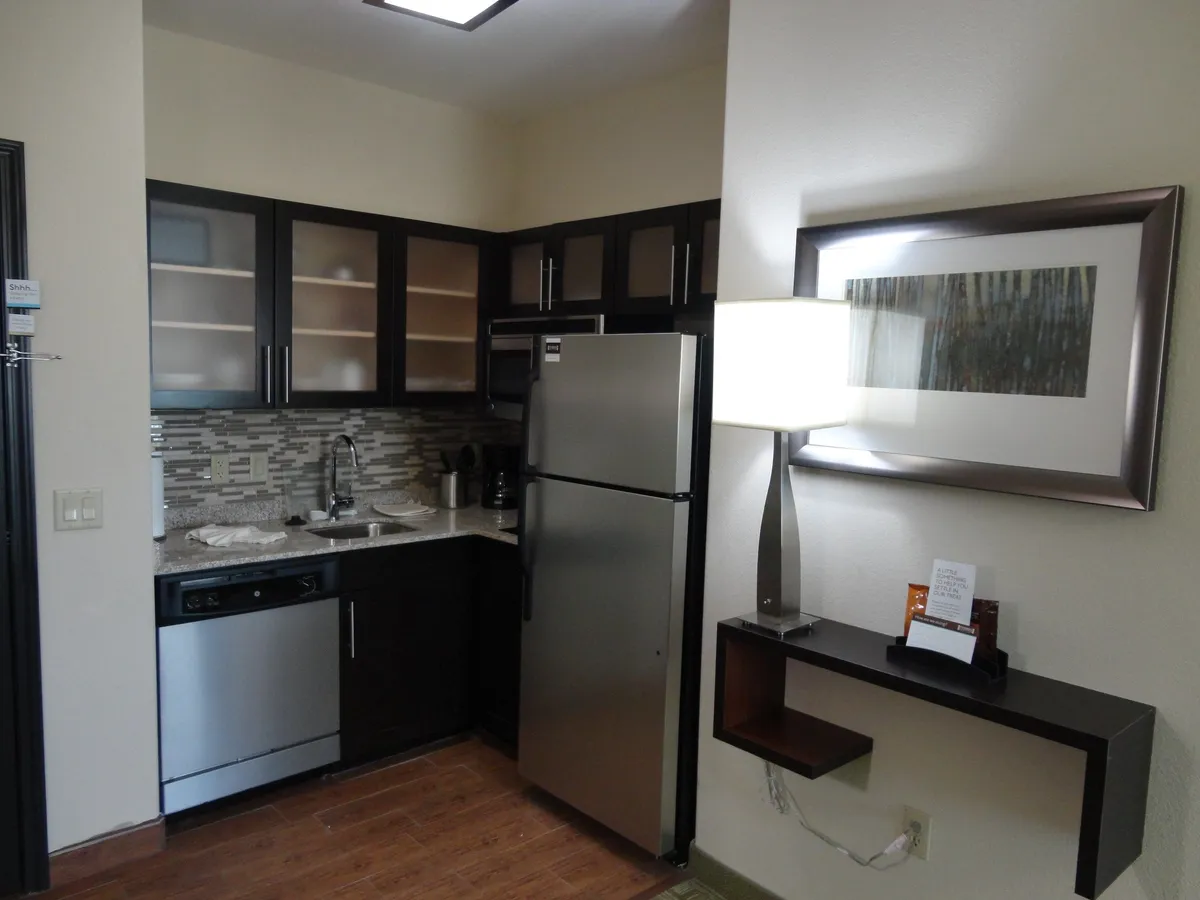 A bright, contemporary in-suite kitchen is on display, featuring modern appliances including a stove, microwave, refrigerator, and dishwasher. The well-lit countertop and overhead lighting create an inviting atmosphere, while tasteful artwork hangs on the wall. The space is set up for cooking, socializing, and enjoying meals together.