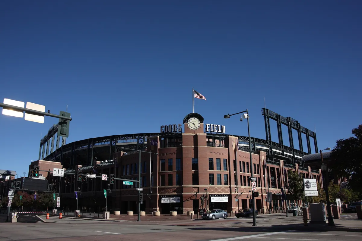 The image captures the iconic exterior of Denver's Coors Field. The scene features the recognizable Coors Field sign atop the stadium, a large clock, the American flag flying proudly, and distinct architectural details that highlight its role as a vibrant sports hub. The energetic atmosphere is palpable, hinting at the excitement of game days and major events.