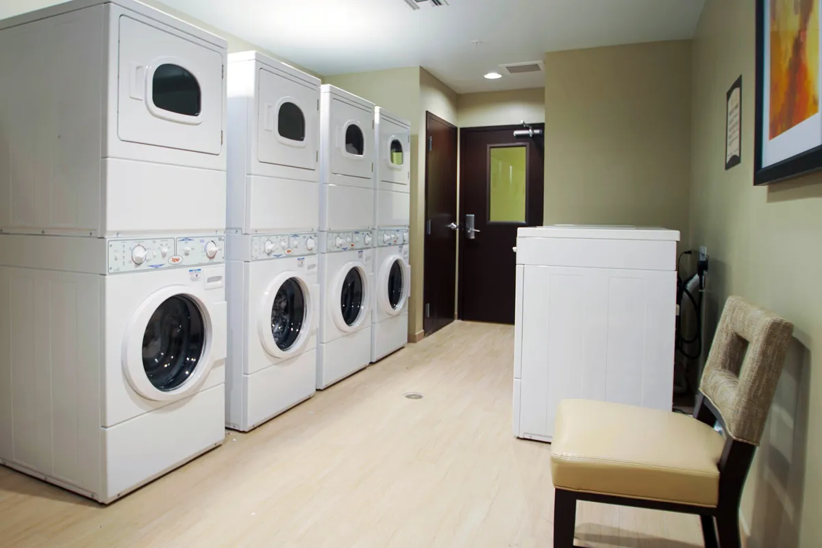 A bright, functional laundry room featuring several stacked washers and dryers along one wall, with a small seating area nearby for guests to wait comfortably. The space is organized and well-lit, emphasizing convenience and practicality.